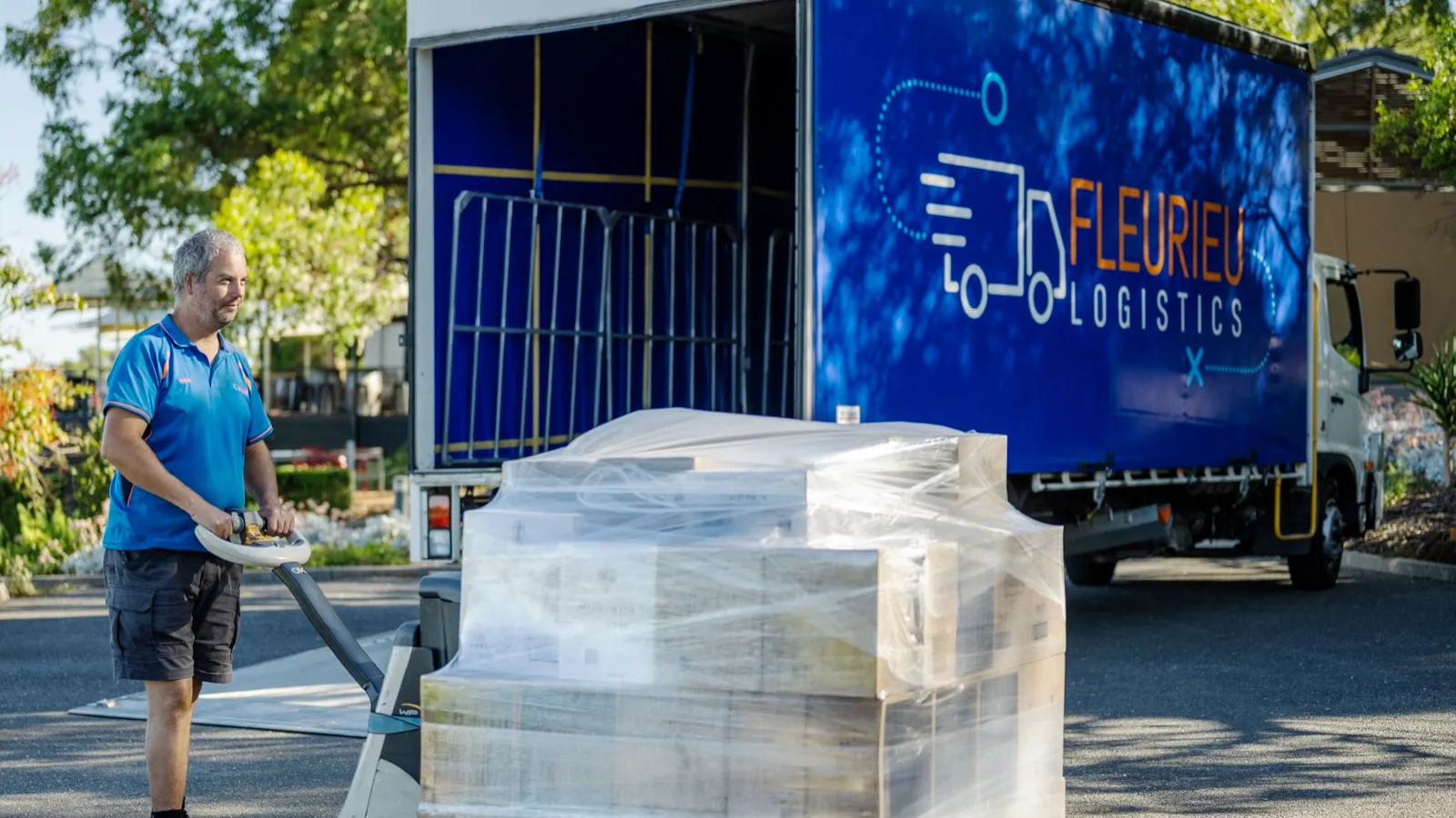 A Fleurieu Logistics driver moving a shrink-wrapped pallet with a pallet jack at ground level, beside one of the fleet's curtain-side trucks with its side curtain open